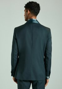 Chaqueta de traje en verde oscuro vista desde la espalda, con un cuello de acabado satinado, hombros estructurados y un corte ajustado con puños abotonados.
