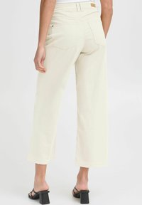 Wide-leg, cream-colored trousers with a high waist, back pockets, and frayed hem. Paired with black block-heel sandals.
