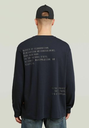 Man met een zwarte pet en een donker shirt met lange mouwen met witte tekst op de rug, staand tegen een effen lichte achtergrond.