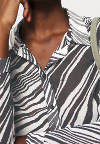 Persona che indossa una camicia a righe zebra in bianco e nero con colletto, appoggiando la mano vicino al viso, porta una borsa a tracolla beige.