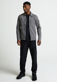 Pelēks jaka ar apkakli, diviem krūšu kabatām un rāvējslēdzēju; apvienota ar melniem kravas biksēm un melniem sporta apaviem. Vienkārša fona.