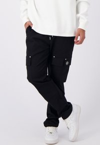 Zwarte cargobroek met meerdere zakken, een elastische taille met trekkoord, en details van zilverkleurig metaal, gecombineerd met witte sneakers en een witte textuurtop.