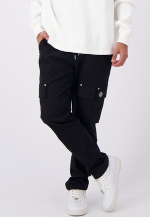 Zwarte cargobroek met meerdere zakken, een elastische taille met trekkoord, en details van zilverkleurig metaal, gecombineerd met witte sneakers en een witte textuurtop.