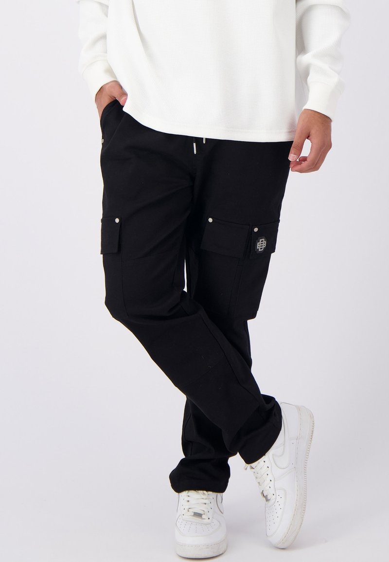 Zwarte cargobroek met meerdere zakken, een elastische taille met trekkoord, en details van zilverkleurig metaal, gecombineerd met witte sneakers en een witte textuurtop.