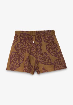 Joven mujer con camisa marrón estampada abotonada anudada a la cintura y pantalones cortos a juego con sandalias negras de punta abierta, de pie frente a un fondo liso.