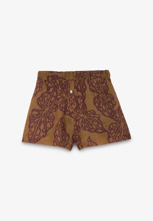 Joven mujer con camisa marrón estampada abotonada anudada a la cintura y pantalones cortos a juego con sandalias negras de punta abierta, de pie frente a un fondo liso.