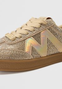 Sneaker dorato con decorazioni in strass, tomaia in tessuto, lacci beige e suola in gomma. Presenta un accento logo iridescente di lato.