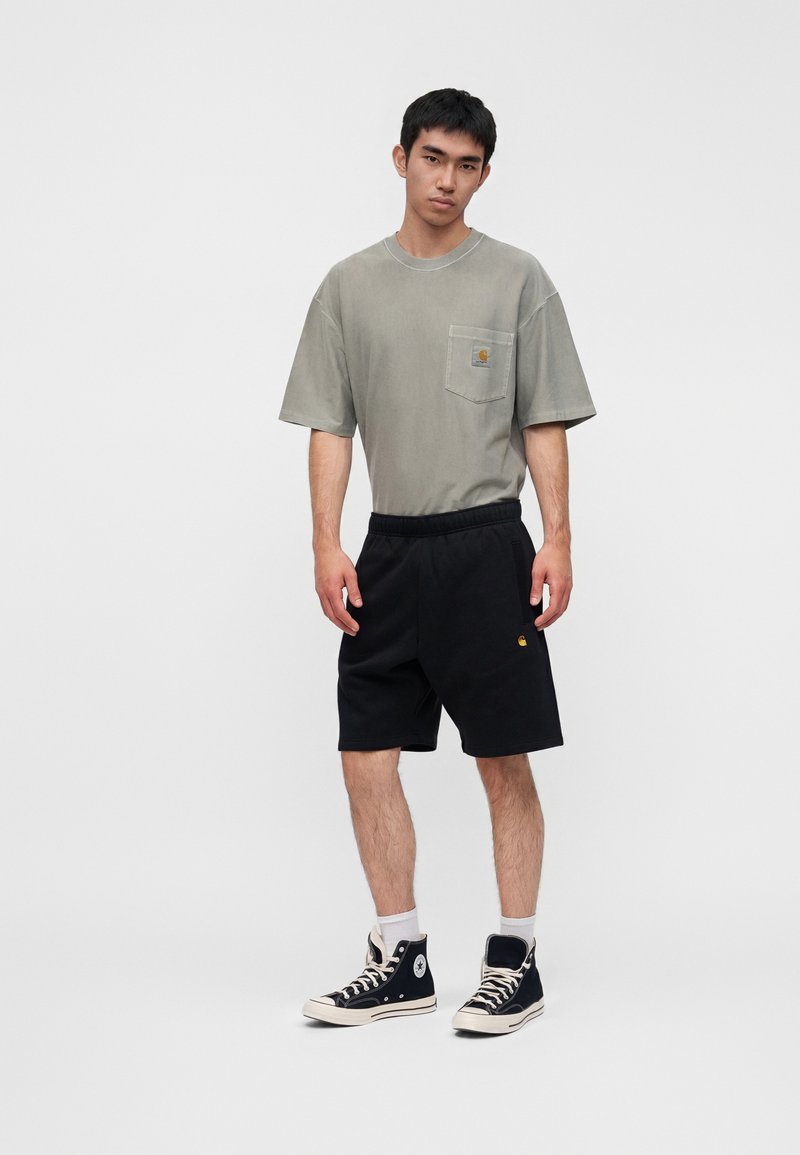 Junger Mann trägt ein ausgewaschenes graues oversized T-Shirt mit Tasche, schwarze Shorts, weiße Socken und schwarze hohe Sneakers und steht vor einem einfarbigen Hintergrund.