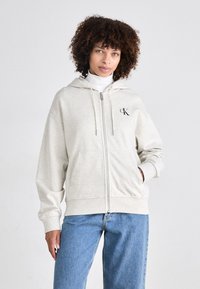 Šviesiai pilkas zip-up gobtuvėlis pagamintas iš minkštos medžiagos, su reguliuojamu gobtuvu, priekinėmis kišenėmis ir mažu juodu logotipu ant krūtinės. Suderintas su mėlynomis džinsais.