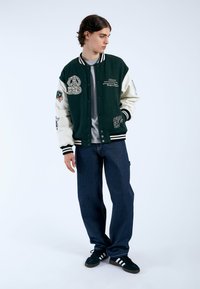 Giacca varsity verde e bianca con toppe ricamate, polsini a costine e vestibilità morbida. Abbinata a pantaloni in denim blu e sneakers nere.