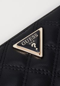 Portafoglio nero in pelle sintetica trapuntata con logo hardware triangolare in tono oro che presenta il testo "GUESS U.S.A." e un dettaglio a punto interrogativo.