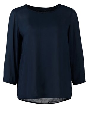 Femme portant un haut bleu marine, une jupe grise, des collants noirs, des bottines noires et tenant une pochette beige.