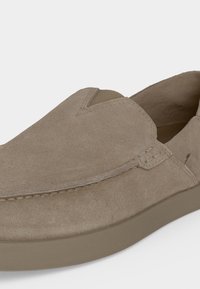 Chaussure slip-on en suède marron clair avec un bout arrondi, des coutures apparentes et une semelle extérieure en caoutchouc plate.