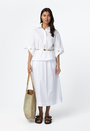 Žena v bielej blúzke so zaväzovaním v páse a sukni, držiaca veľkú pletenú tašku a obutá v čiernych sandáloch s cvočkami, stojaca na bielom pozadí.