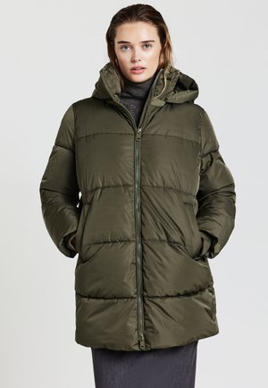 Femme portant une doudoune à capuche vert olive, les mains dans les poches, accompagnée d'un turtleneck sombre et d'une jupe, faisant face à l'avant.