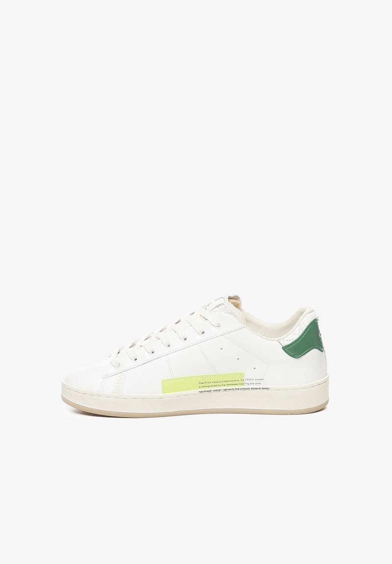 Sneaker bianca in pelle con dettaglio verde sul tallone, suola beige testurizzata e un'etichetta gialla prominente sul lato che presenta un riassunto dell'artigianato.