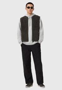 Gilet sombre doublé en polaire sur un sweat à capuche gris clair, associé à un pantalon en velours côtelé noir et des baskets grises. Le mannequin porte des lunettes de soleil noires.