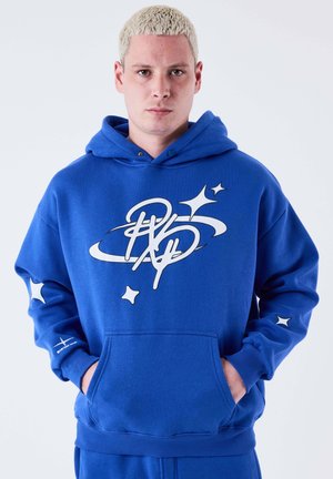 Joven con cabello rubio corto que lleva una sudadera azul con diseños blancos de estrellas y remolinos, manos en el bolsillo delantero, expresión neutra.