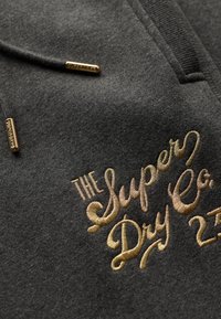 Czarna szara tkanina bluzy z kapturem z złotą haftowaną napisem "THE Super Dry Co." oraz dekoracyjnymi złotymi akcentami.