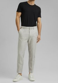 Homme portant une chemise noire à manches courtes, un pantalon slim gris clair et des baskets blanches, debout avec une main dans la poche.