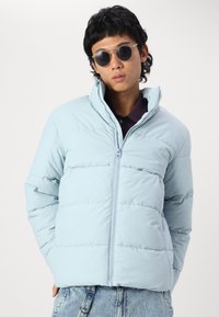 Light blue puffer jacket with a high collar, stitched panels, and a front zipper, worn with jeans and round sunglasses.