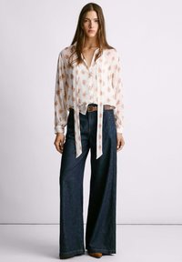 Camicia bianca con motivo floreale rosa e dettaglio annodato, abbinata a pantaloni a gamba larga in denim scuro e cintura marrone. Sono visibili scarpe marroni.