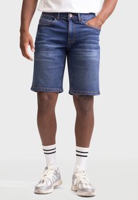 Denimshorts i mørkeblå med falmede områder, fem lommer og kontraststing. Brukt med hvite sokker med svarte striper og sneakers.