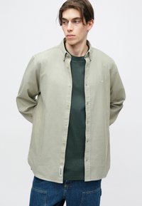 Camisa de botonadura ligera verde sobre una camiseta verde oscuro, combinada con jeans azules. Presenta un diseño simple y una etiqueta en el dobladillo.