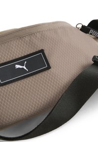 Bolso de cintura Puma en malla beige con cierre de cremallera y correa negra. Presenta un parche cuadrado negro con logo blanco. Superficie texturizada.