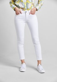 Weiße, enge Jeans aus einem glatten Material, mit Knopf- und Reißverschluss, seitlichen Taschen und einer taillierten Passform. Kombiniert mit weißen Sneakers.