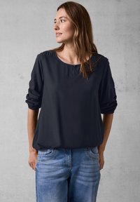 Mørkeblå, langærmet bluse med en afslappet pasform, der har en rund halsudskæring og en knapdetalje foran. Sammen med lyseblå jeans.