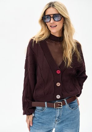 Bordeauxrode gebreide cardigan met kabelpatroon en drie knopen in roze, crème en beige. Gecombineerd met lichtblauwe spijkerbroek en een bruine riem.