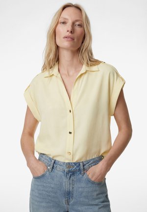 Blondynka z rękami w kieszeniach, ubrana w bluzkę z krótkim rękawem w jasnożółtym kolorze z guzikami oraz jasnoniebieskie dżinsy z wysokim stanem na białym tle.