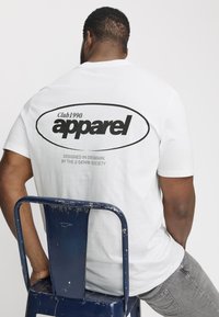 Camiseta de algodón blanca con un gran gráfico de texto negro en la parte posterior, que dice "Club1990 apparel", con detalles de diseño adicionales a continuación.
