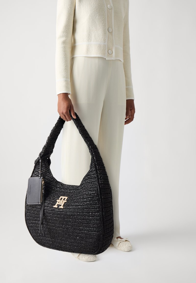 Personne portant une tenue crème tenant un grand sac à main tressé noir avec un logo doré et une pochette noire attachée sur fond uni.