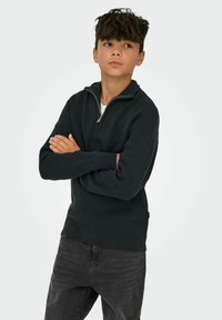 Pull en maille côtelée vert foncé avec un col zippé à mi-hauteur, comportant des manches longues, porté par-dessus un t-shirt blanc, associé à un jean foncé.