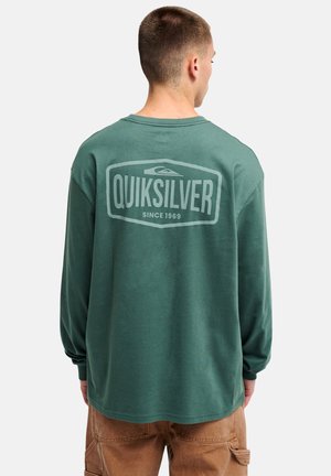 Man wearing green long-sleeve shirt with "Quiksilver Since 1969" logo on back, paired with brown pants, standing facing away.