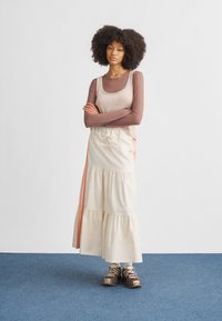 Kevyt, kerroksittain levenevä beige hame, jossa on joustava vyötärönauha ja oranssit sivuraidat. Yhdistetty pitkähihainen ruskea paita ja rennot tennarit.