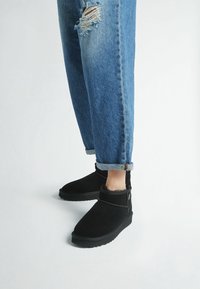 Botines de ante negros con un suave forro de piel, suela de goma plana y una correa decorativa en la parte frontal, combinados con unos vaqueros azules arremangados.