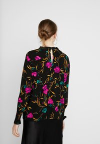 Schwarze Bluse mit langen Ärmeln, die ein florales Muster in Pink, Blaugrün und Gold zeigt, sowie ein Schlüssellochdetail im Rückenteil. Texturstoff.
