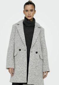 Manteau texture gris clair avec de grands revers, boutons croisés et poches latérales, superposé à un pull à col roulé noir.
