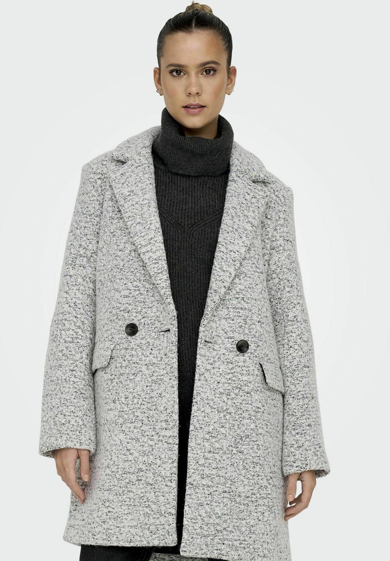 Manteau texture gris clair avec de grands revers, boutons croisés et poches latérales, superposé à un pull à col roulé noir.
