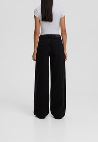 Jeans negros de pierna ancha con cintura alta, tejido de mezclilla, grandes bolsillos traseros y un detalle de costura mínimo, combinados con una camiseta gris ajustada.