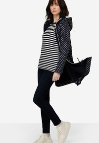 Merihenkinen takki, jossa on puku ja huppu, raidallisen paidan päällä, yhdistettynä tummiin farkkuihin ja valkoisiin tennareihin, esitellen rentoa kerroksellista tyyliä.