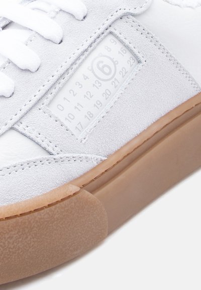 Baskets blanches en daim et cuir, avec une semelle en caoutchouc, des lacets plats blancs et une étiquette carrée affichant des chiffres et un logo.