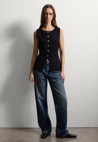 Schwarzes ärmelloses Oberteil mit goldenen Knöpfen an der Vorderseite, kombiniert mit weiten blauen Jeans. Glattes Material, strukturierte Form und minimalistisches Design.