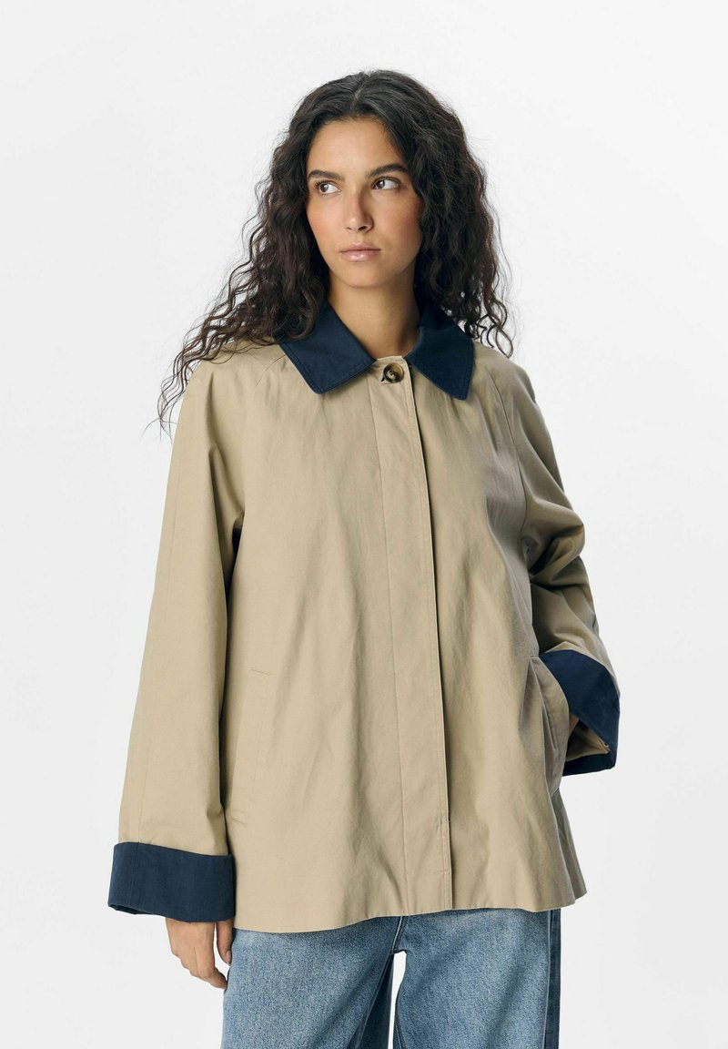 Beige jasje met marineblauwe kraag en manchetten, voorzien van een knoopsluiting en zijzakken. Gemaakt van een glad, lichtgewicht materiaal.