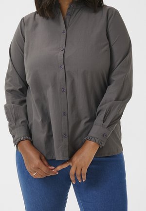 Femme portant une blouse boutonnée gris foncé avec des poignets à volants et un jean bleu, les mains légèrement jointes à la taille.