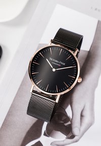 Montre noire avec un cadran rond noir, des accents en or rose et un bracelet en maille. Présente des marqueurs d'heure simples et un design minimaliste.