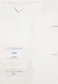 Interno di un indumento bianco con etichette che leggono "MNG" e "SLIM FIT". Presenta un design pulito con cuciture a vista e dettagli dei fori dei bottoni.